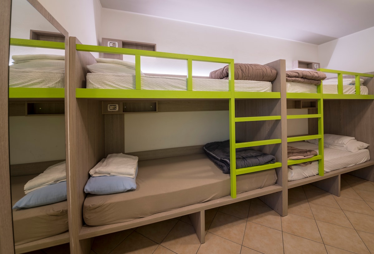 A room with three sets of bunk beds is presented, featuring warm wood finishes and green accents. Each bed is fitted with a mattress and accompanied by a shelf for personal items. Reading lights and power outlets are available at each sleeping area.