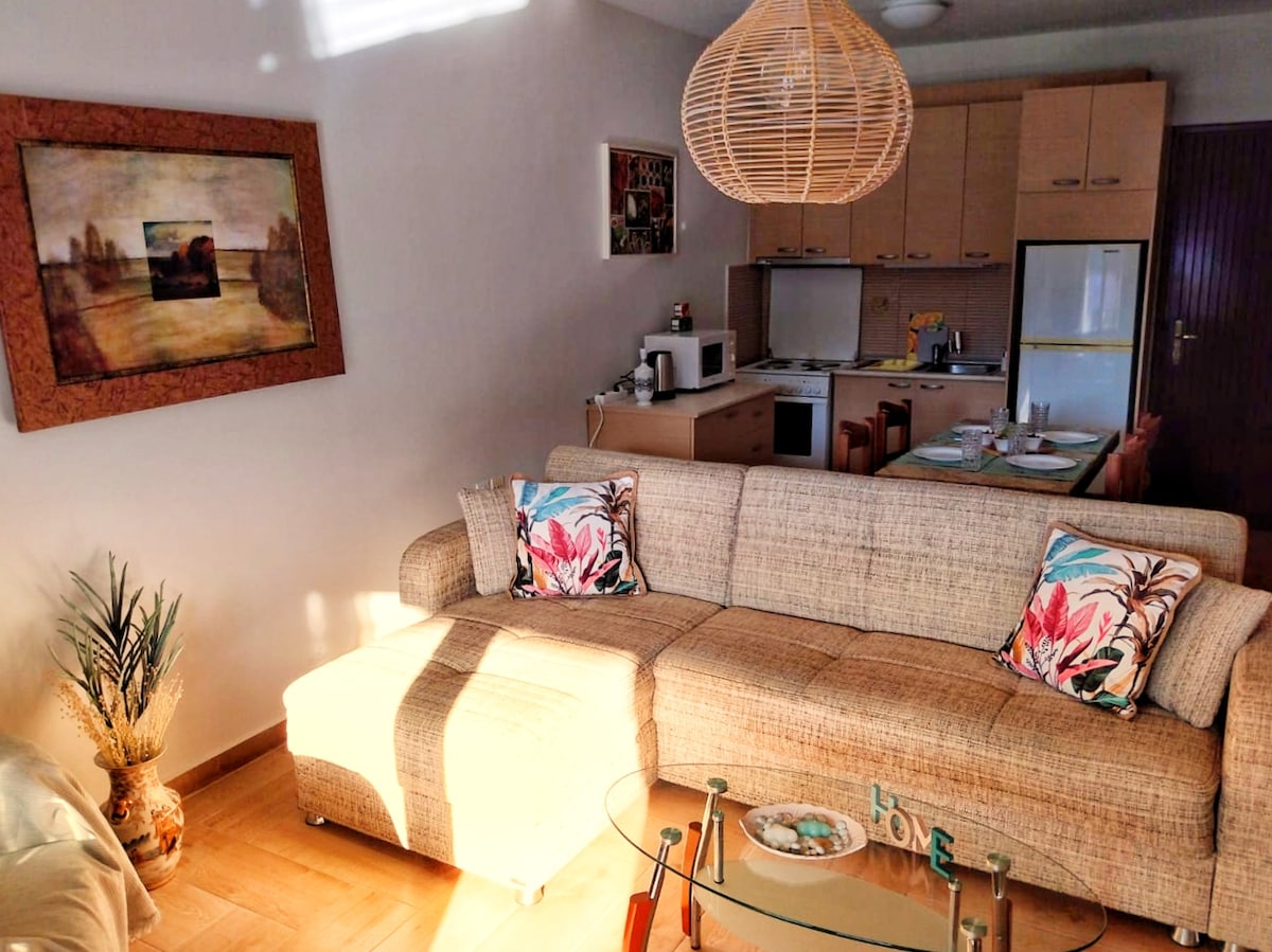 A well-lit living area features a comfortable couch adorned with patterned cushions. A glass coffee table sits in front of the couch, and a wooden dining table is visible in the background. The space includes a kitchenette, highlighted by a mounted microwave and a refrigerator, with artwork decorating the walls.