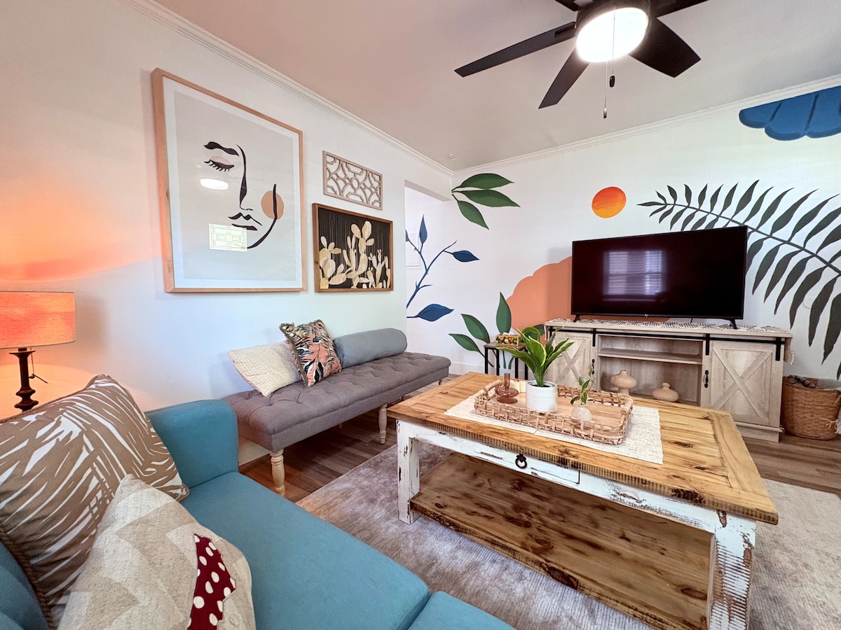 A living area features a mix of seating options, including a blue sofa and a gray sofa. A rustic coffee table sits in the center, complemented by decorative items. Colorful wall murals, including botanical and abstract elements, create a vibrant atmosphere, while a television is positioned on a light wooden console.