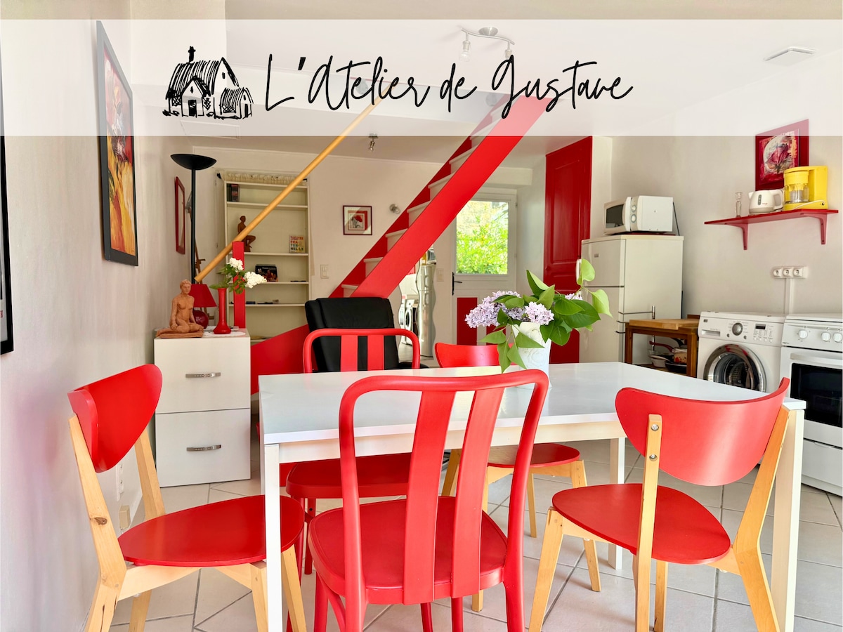 A bright dining area is highlighted by a white table surrounded by red chairs. The open space leads to a cozy kitchen, with artwork adorning the walls. A staircase in the background connects the two levels, and natural light brightens the room.