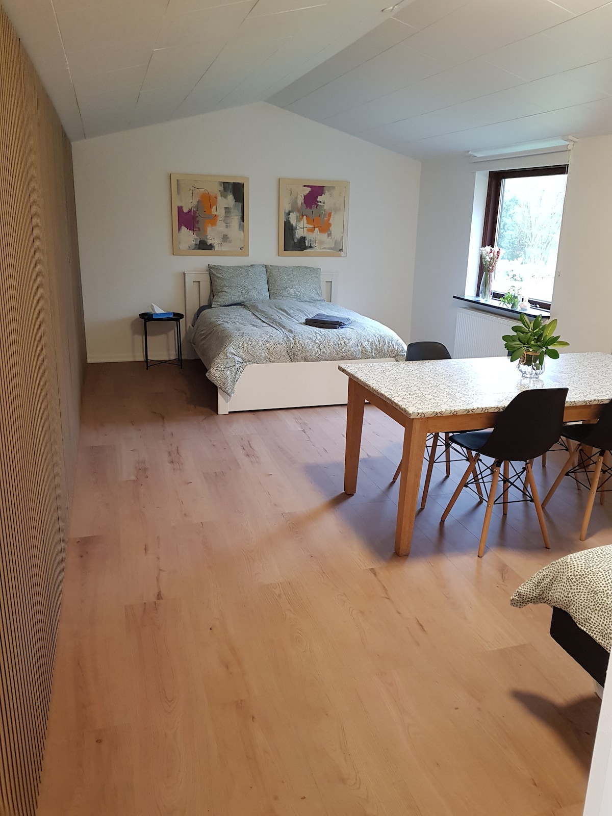 A spacious room features a comfortable bed with a light-colored quilt and two abstract artwork pieces hung above. A dining table with black chairs is positioned beside the bed, while a window provides natural light, enhancing the overall ambience of the space.