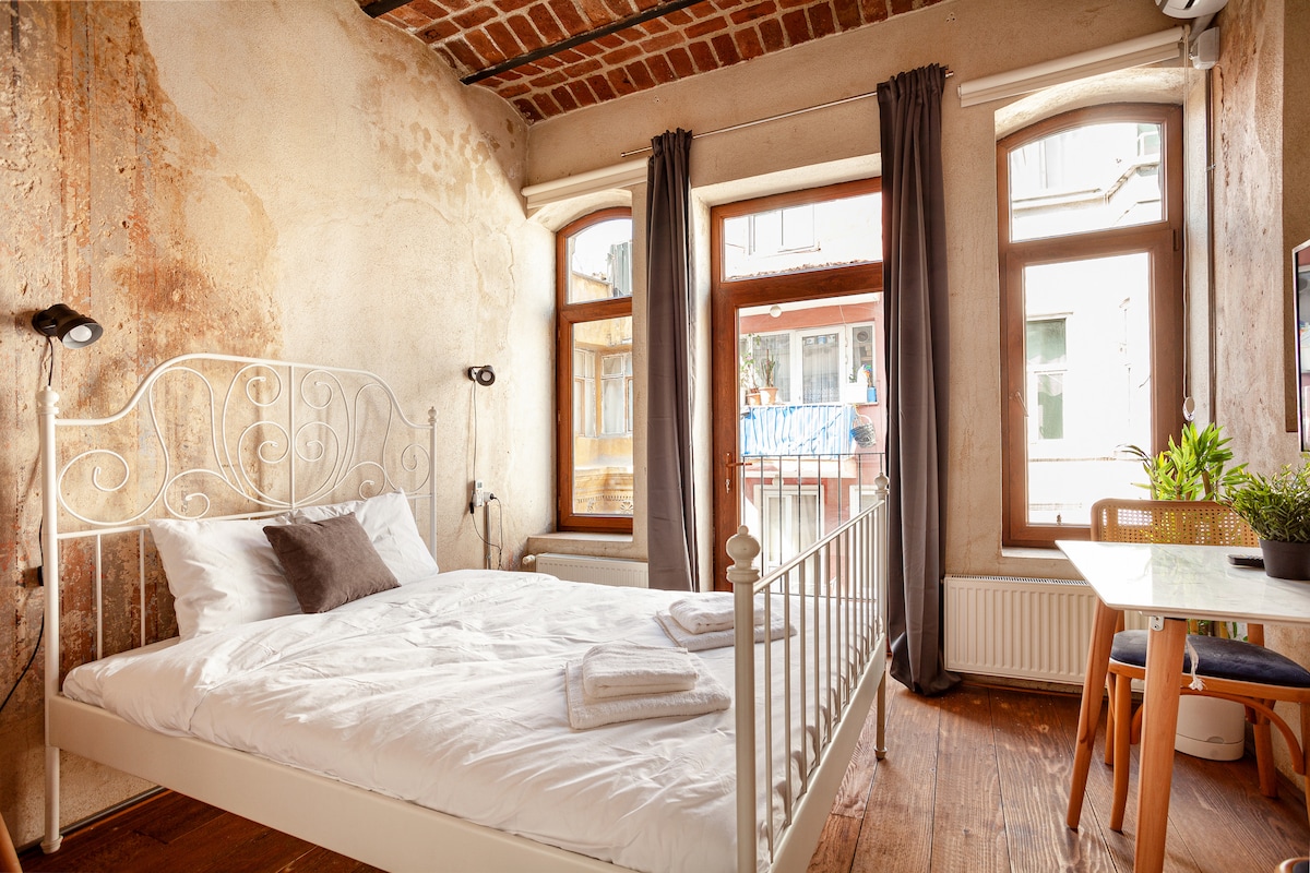 A well-lit bedroom features a spacious white bed with neatly arranged linens. Two large windows frame the room, with light filtering through dark curtains. A small, wooden table is present alongside a plant, enhancing the inviting atmosphere of the space.