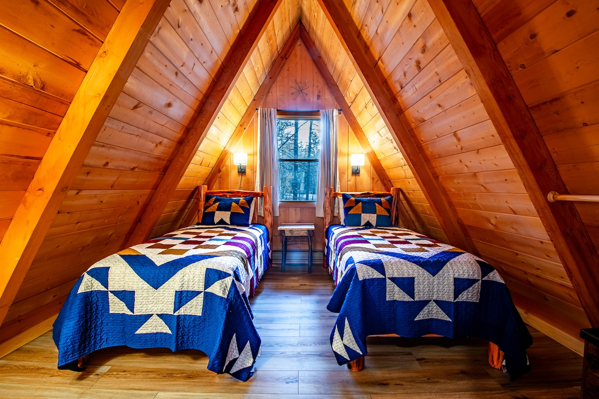 An upstairs bedroom features two twin beds adorned with blue and white quilt patterns. Natural light enters through a window set within the A-frame structure, illuminating the warm wood paneling and creating a cozy atmosphere. A small bedside table is positioned between the beds.