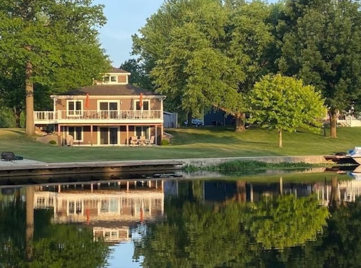 A spacious lake house is depicted, featuring a large outdoor deck and multiple windows that reflect in the calm waters. Surrounding trees provide greenery, while the well-kept yard offers areas for relaxation. The private dock is visible, adding to the serene lakefront setting.