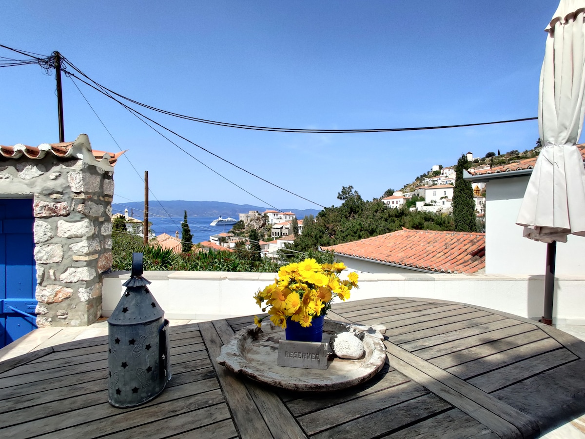 A wooden table is set on a terrace, featuring a blue pot filled with bright yellow flowers. The background showcases a sea view with rooftops of traditional houses against a clear blue sky.