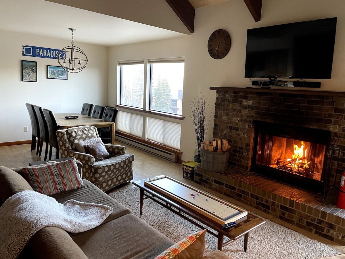 A warm living area is illuminated by a wood-burning fireplace, featuring a comfortable sofa and patterned armchair. A wooden coffee table is positioned in front of the fireplace, and a dining table for six is visible nearby. Large windows provide natural light and views of the surrounding landscape.