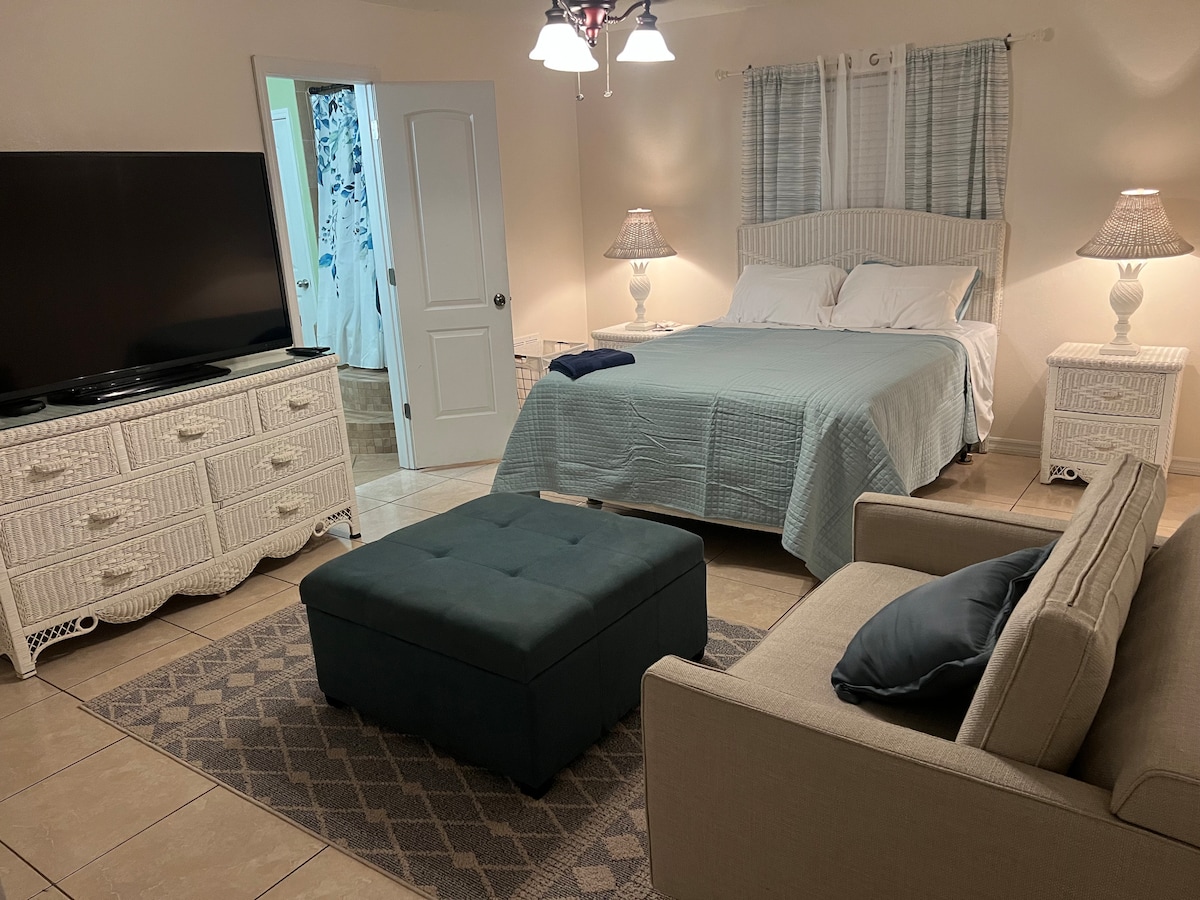 A spacious bedroom is shown, featuring a Queen-sized bed with a light blue quilt. Two bedside tables with lamps provide illumination, and a television sits on a white dresser. An ottoman in the center and a cozy armchair complete the inviting space.