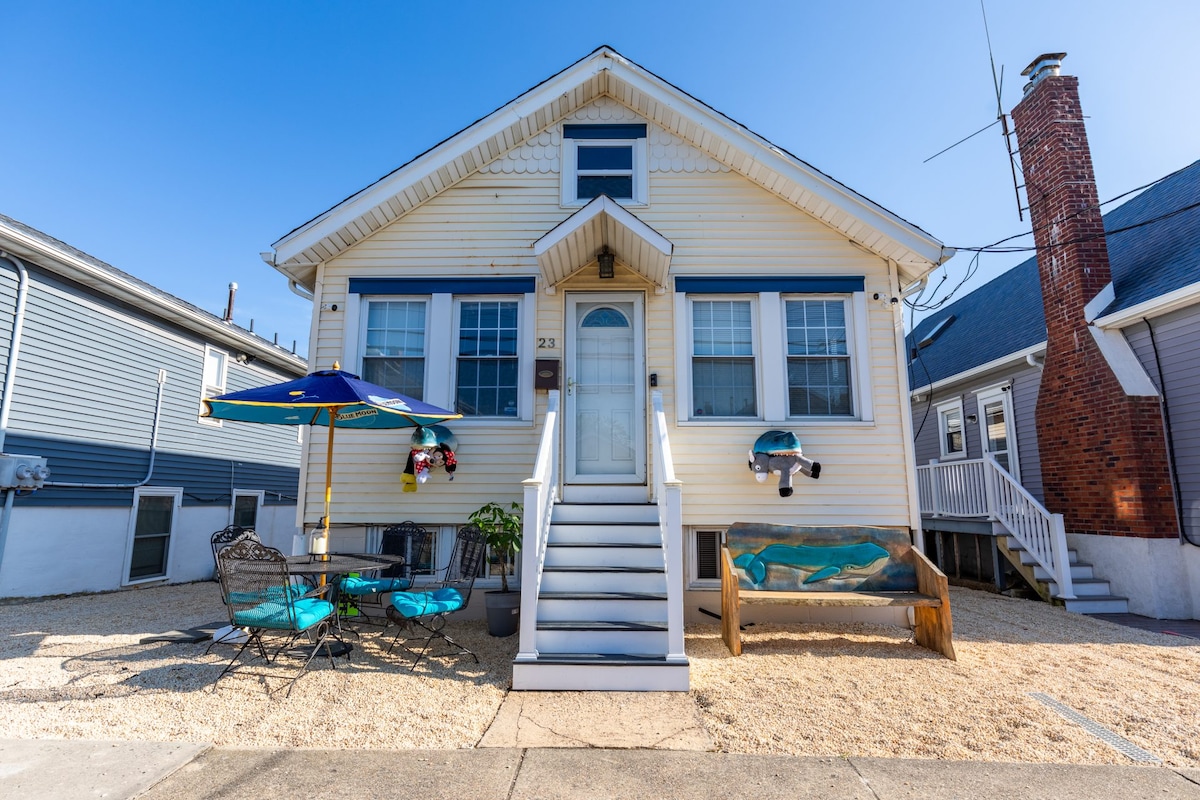 A charming two-story house is presented, featuring a welcoming front porch and a well-maintained facade. Outdoor seating is arranged at a round table shaded by a colorful umbrella. A decorative bench sits beside the entrance, and light blue skies complete the inviting exterior.