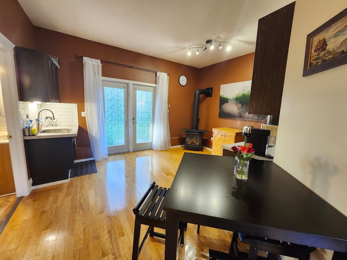 An open living space features a dining table with a vase of flowers. A gas fireplace adds warmth, while large glass doors allow natural light and provide access to the exterior. The kitchen area is visible to the left, showcasing cabinetry and a counter.