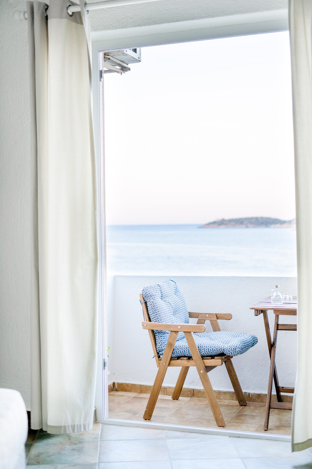A cozy balcony space is presented with a wooden chair and table, both positioned to enjoy the sea view. Light, airy curtains frame the open doorway, while the tranquil turquoise water is visible in the distance, offering a serene atmosphere.