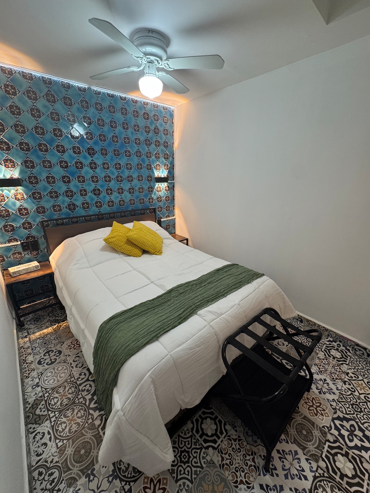 A comfortable sleeping area is presented with a spacious bed, adorned with soft bedding and two decorative pillows. A fan is mounted on the ceiling, adding to the room's ventilation. Unique patterned tiles cover the floor beneath a feature wall of blue and patterned tiles.