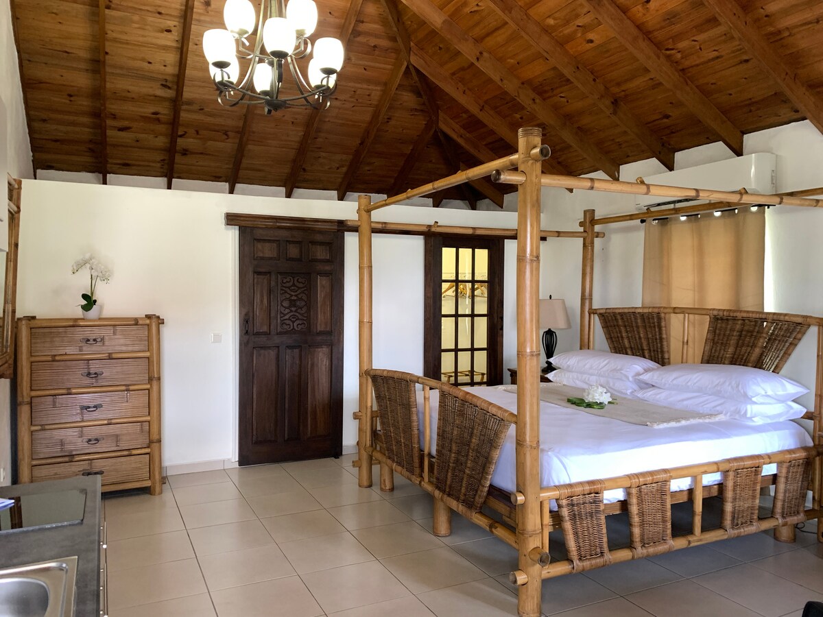A spacious guest room features a bamboo four-poster bed adorned with crisp white linens. Natural wood accents highlight the ceiling and furniture. A wooden dresser stands against one wall, while a small kitchenette is visible in the foreground, contributing to the room's functional design.