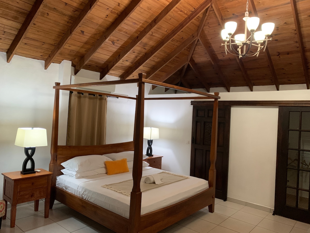 A spacious guest room features a wooden four-poster bed with white linens and an accent pillow. Two bedside tables with lamps provide soft lighting. The ceiling showcases exposed wooden beams, and a chandelier adds illumination. Double doors lead to an inviting outdoor area.