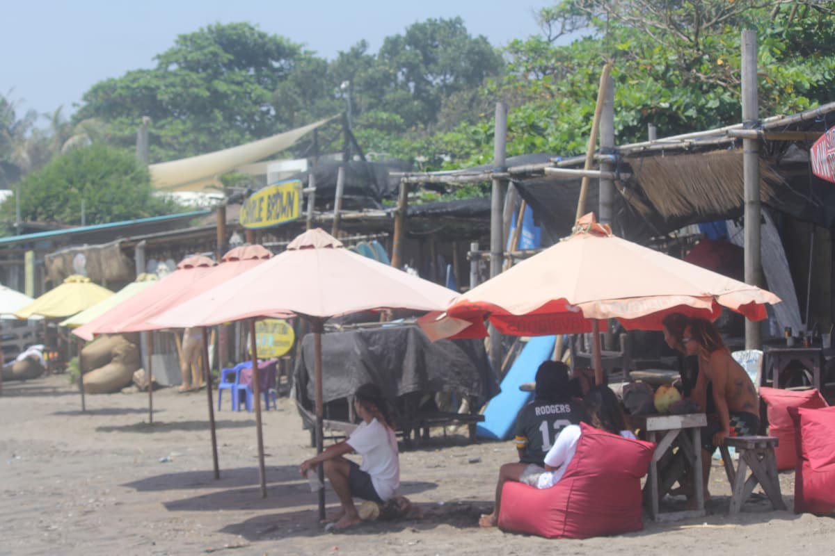 Shaded seating areas with colorful umbrellas provide respite along the beach. The sandy ground is dotted with beanbag chairs, while simple wooden tables are seen beneath the umbrellas. A rustic ambiance is created by nearby thatched structures and foliage in the background.