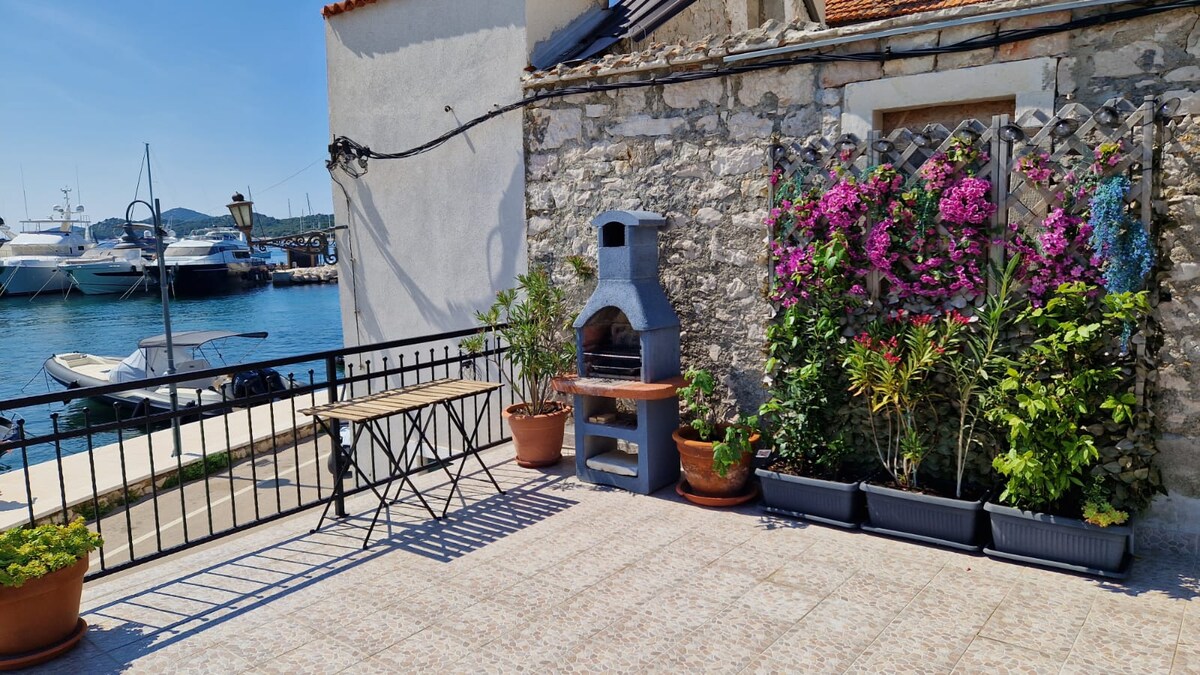 An outdoor terrace is presented with a stone wall backdrop, complemented by vibrant flowers in pots and a charcoal grill. A wooden table and folding chairs are positioned for dining, while the view of the harbor and boats adds a tranquil setting.
