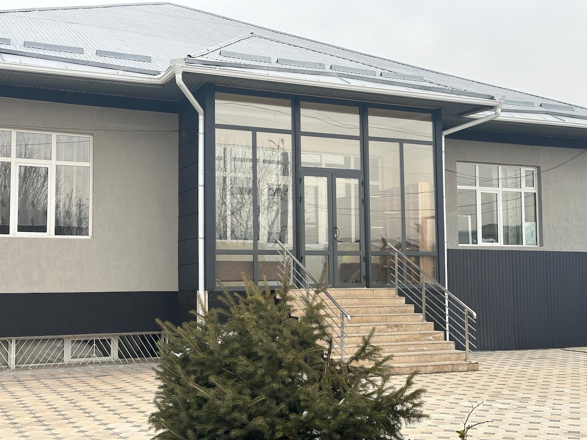 The entrance of the hostel is shown, featuring a set of wide stairs leading to a glass door. Large windows flank both sides of the entrance, providing an open and welcoming appearance. The ground is paved with patterned tiles, and a shrub is visible in the foreground.