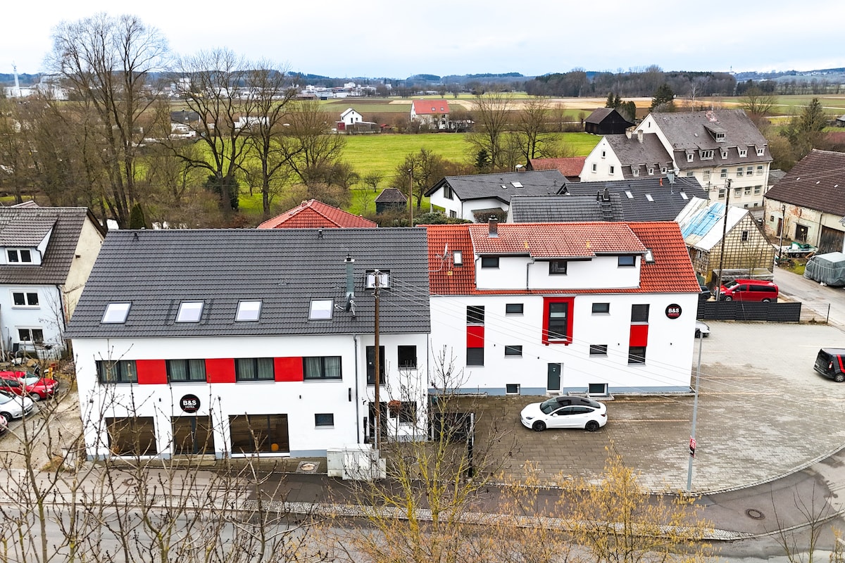 An aerial view of the modern building complex showcases a mix of two structures, one with a dark roof and red accents, alongside a white building. Surrounding green fields and neighboring houses create a serene rural atmosphere.
