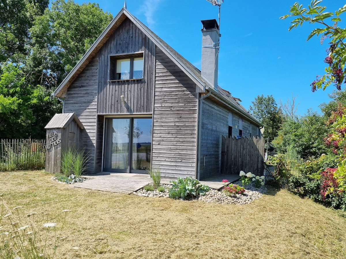 The exterior of the villa showcases a wooden façade and large glass doors opening onto a wooden deck. A grassy area surrounds the home, with plants and decorative stones adding natural elements. A sky filled with blue offers a bright backdrop.