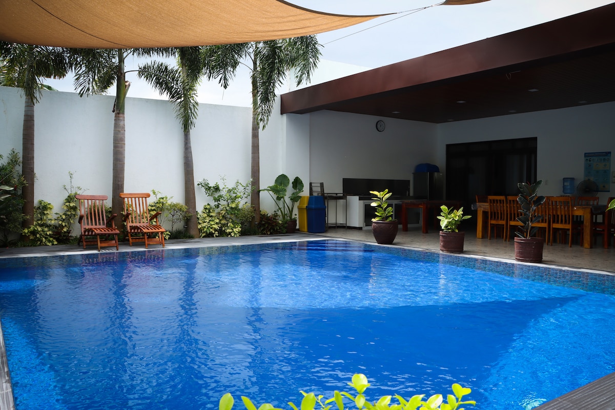 The spacious outdoor area features a large pool with clear blue water, surrounded by tropical plants and palm trees. Several wooden lounge chairs are positioned nearby, and a shaded patio area with dining tables can be seen in the background, offering a relaxing retreat.