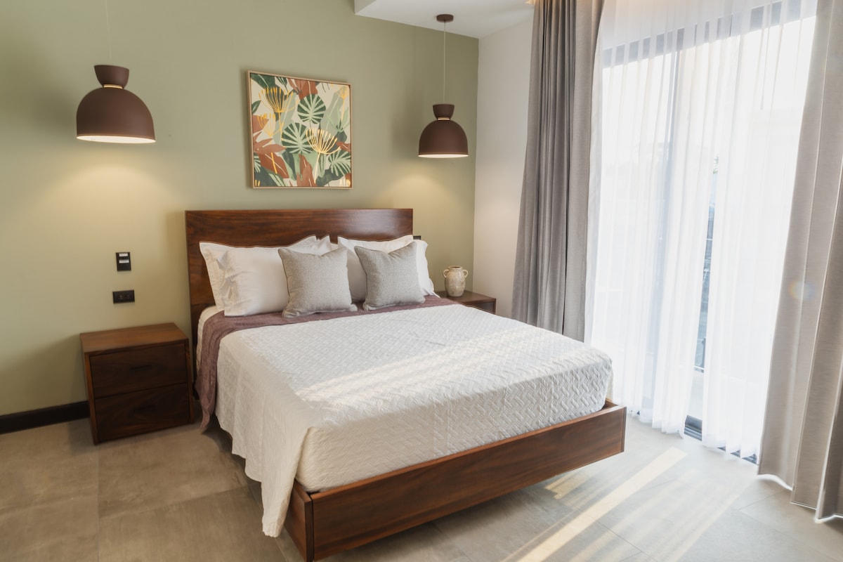 A spacious sleeping area features a wooden bed frame with a light-colored quilt. Two pendant lights hang above the bed, and a decorative wall art piece adds a splash of color. Sheer curtains allow natural light to filter in from a sliding door.