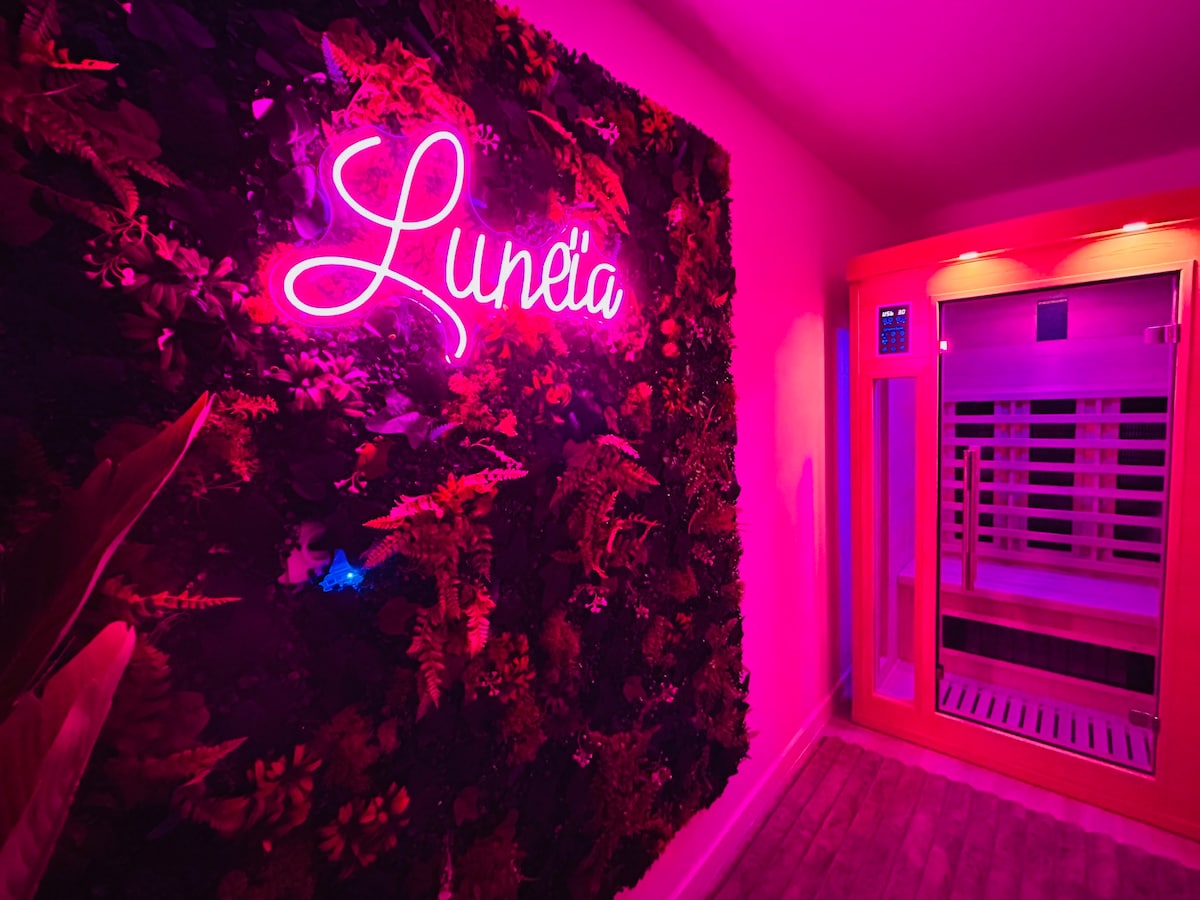 A vibrant wall adorned with lush greenery features the illuminated sign 'Luneïa' in a soft pink hue. Adjacent to it, a modern sauna is visible, showcasing wooden slats and a control panel, all set against a warm, inviting backdrop.