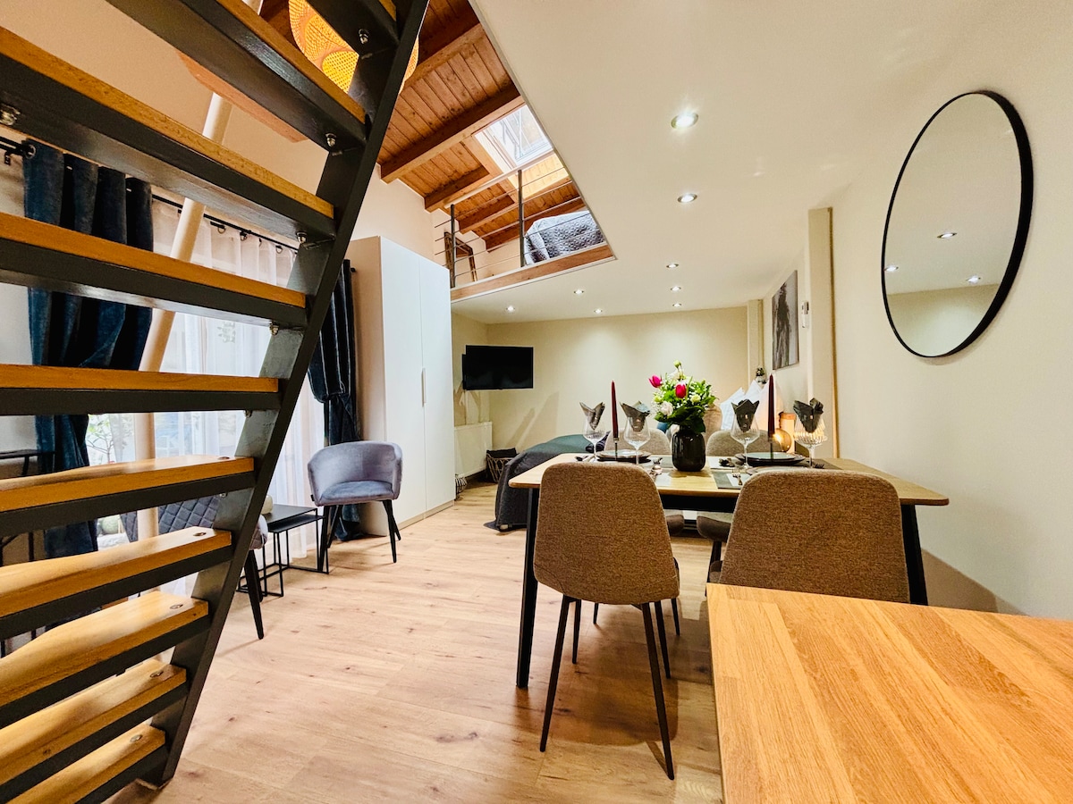 A spacious interior features an open-plan living area with a dining table surrounded by four chairs. A modern staircase leads to the upper level, while a large skylight allows natural light to brighten the space. Decorative elements include a floral centerpiece and stylish seating arrangements.