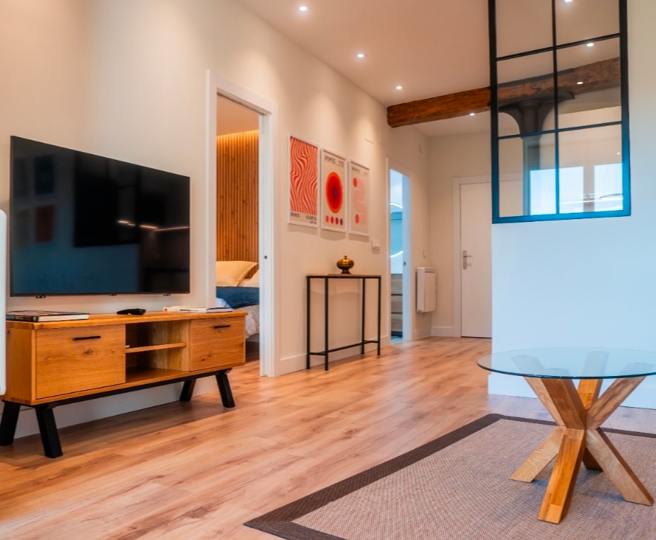 A modern living space features a wooden coffee table and a stylish TV unit with a flat-screen television. Light wood flooring runs throughout, complemented by a decorative area rug. Wall art adds a vibrant touch, and natural light enters through a glass partition, enhancing the airy atmosphere.