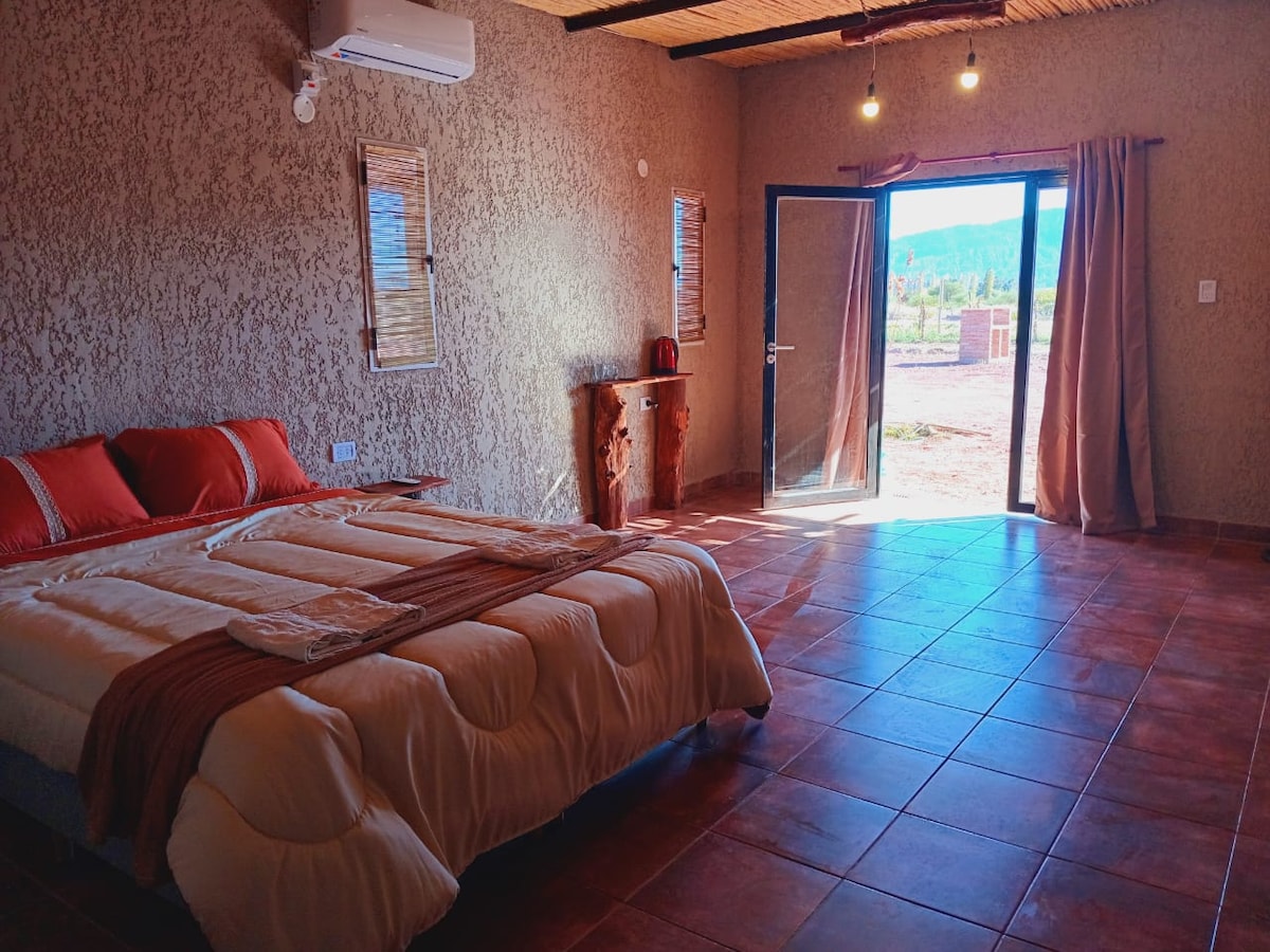 A spacious room features a large bed with cream-colored bedding and coordinating pillows. Natural light floods in through the door leading to an outdoor view of rolling hills. A wooden side table and air conditioning unit are also present, complementing the earthy wall texture.