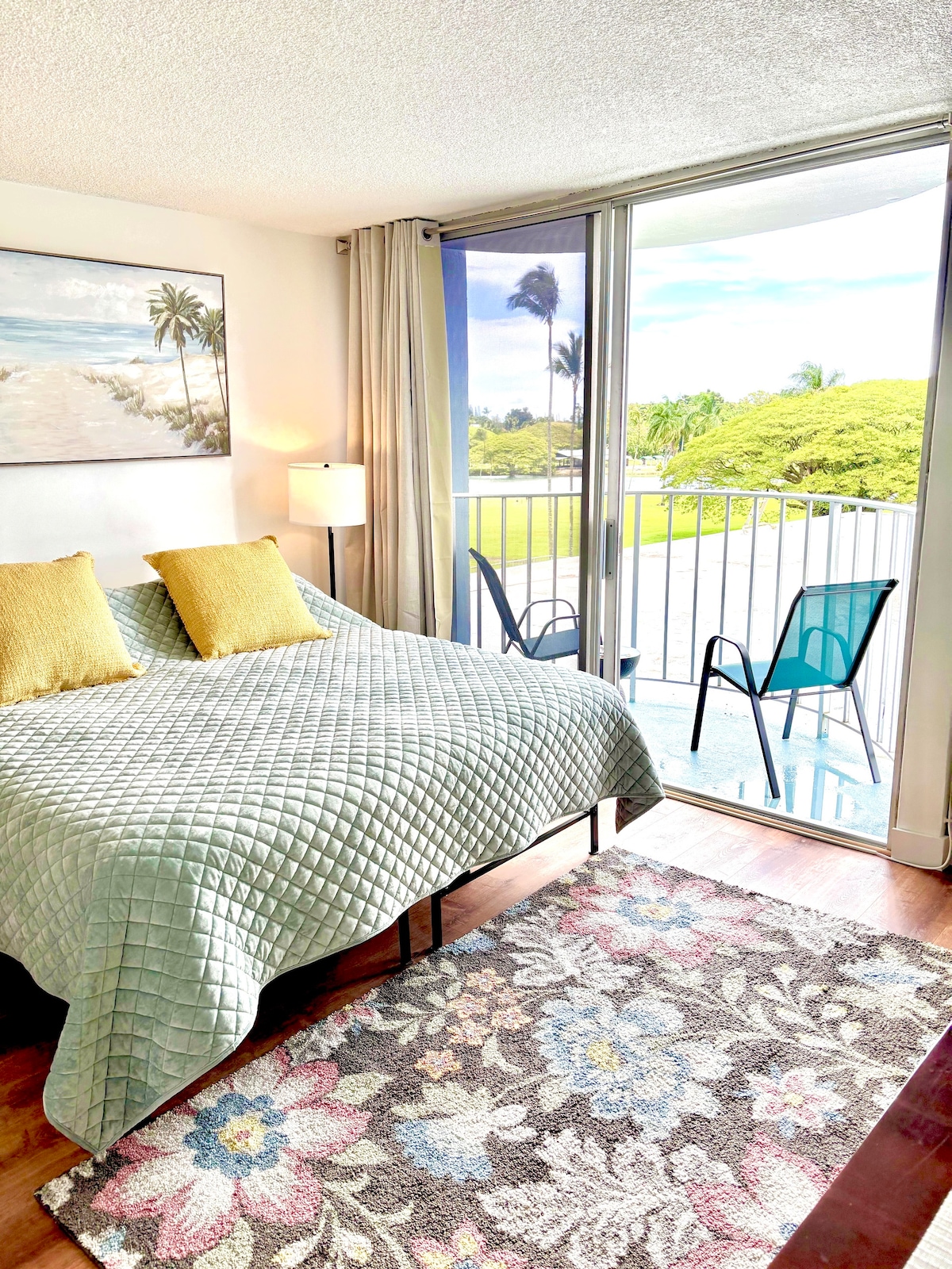 A cozy sleeping area features a spacious bed with a soft quilt, accompanied by two yellow pillows. A large window invites natural light, revealing a view of lush greenery and palm trees. A colorful area rug adds warmth, and a small balcony with blue chairs is visible.