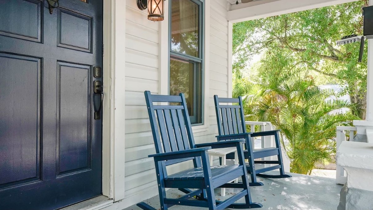 Two blue rocking chairs are positioned on a porch in front of a dark front door. Natural light filters in through the surrounding greenery, creating a relaxed atmosphere. A lantern hangs beside the door, adding a welcoming touch to the entrance.