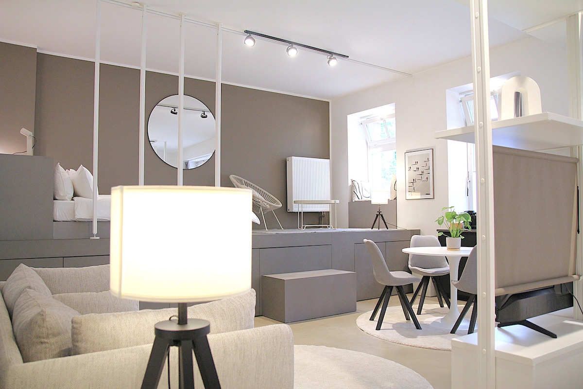A modern living space is showcased with a neutral color palette. Cozy sofas are positioned around a circular rug, complemented by a circular mirror on the wall. Two windows allow natural light to fill the area, while a small dining table and chairs complete the arrangement.