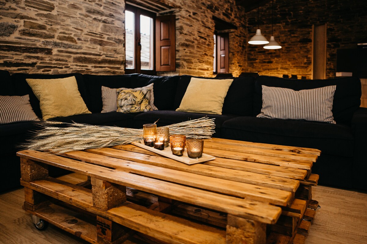 A spacious living area features a large dark sofa adorned with patterned pillows. A rustic wooden coffee table made of pallets holds decorative candles and natural decor. Stone walls and soft lighting create a relaxed ambiance while wooden windows provide natural light.