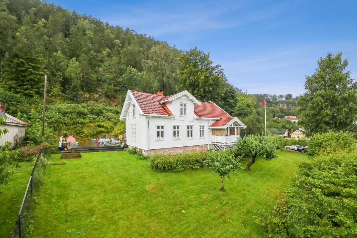 A charming two-story house is nestled in a lush green landscape, surrounded by trees and gardens. The well-maintained exterior features a red-tiled roof and white walls. Open spaces are visible on the property, offering a sense of tranquility and openness.