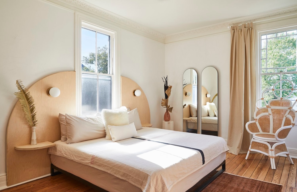 A bright sleeping area features a spacious bed with a light-colored duvet. Natural light enters through a large window, illuminating the minimalist decor. Reflective mirrors and a wooden accent create a serene atmosphere, complemented by decorative elements and a chair positioned nearby.