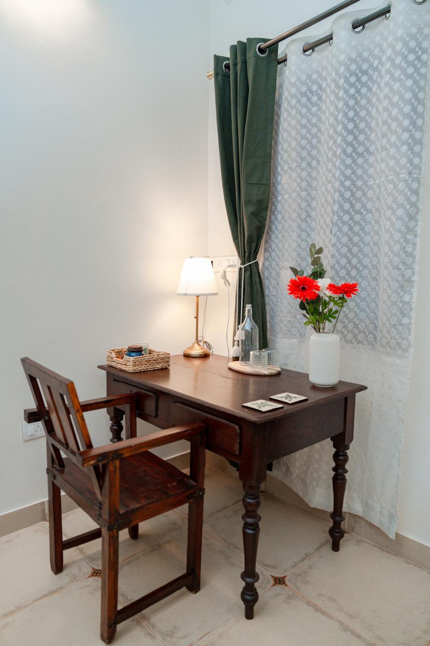 A small wooden desk is positioned against a wall, accompanied by a matching wooden chair. A lamp with a soft glow sits on the desk, alongside a bottle of water and a small vase with vibrant red flowers. Sheer white curtains gently frame the scene.