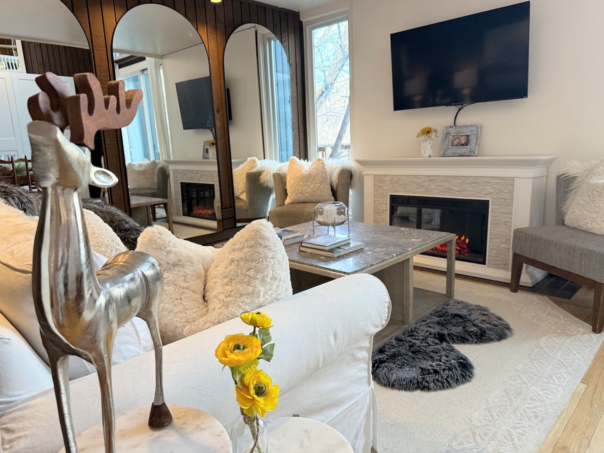 A welcoming living area features soft, textured furnishings, including a cozy sofa and armchairs. A modern fireplace is positioned beneath a large television, providing a focal point. Decorative elements include a sculpture and flowers, enhancing the room's comfort and inviting nature.