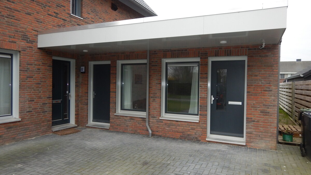 The exterior of the guesthouse is characterized by a brick façade and a covered entrance. Three doors are positioned alongside large windows, allowing natural light to enter. A paved area in front complements the modern design, which is aesthetically balanced with the surrounding greenery.
