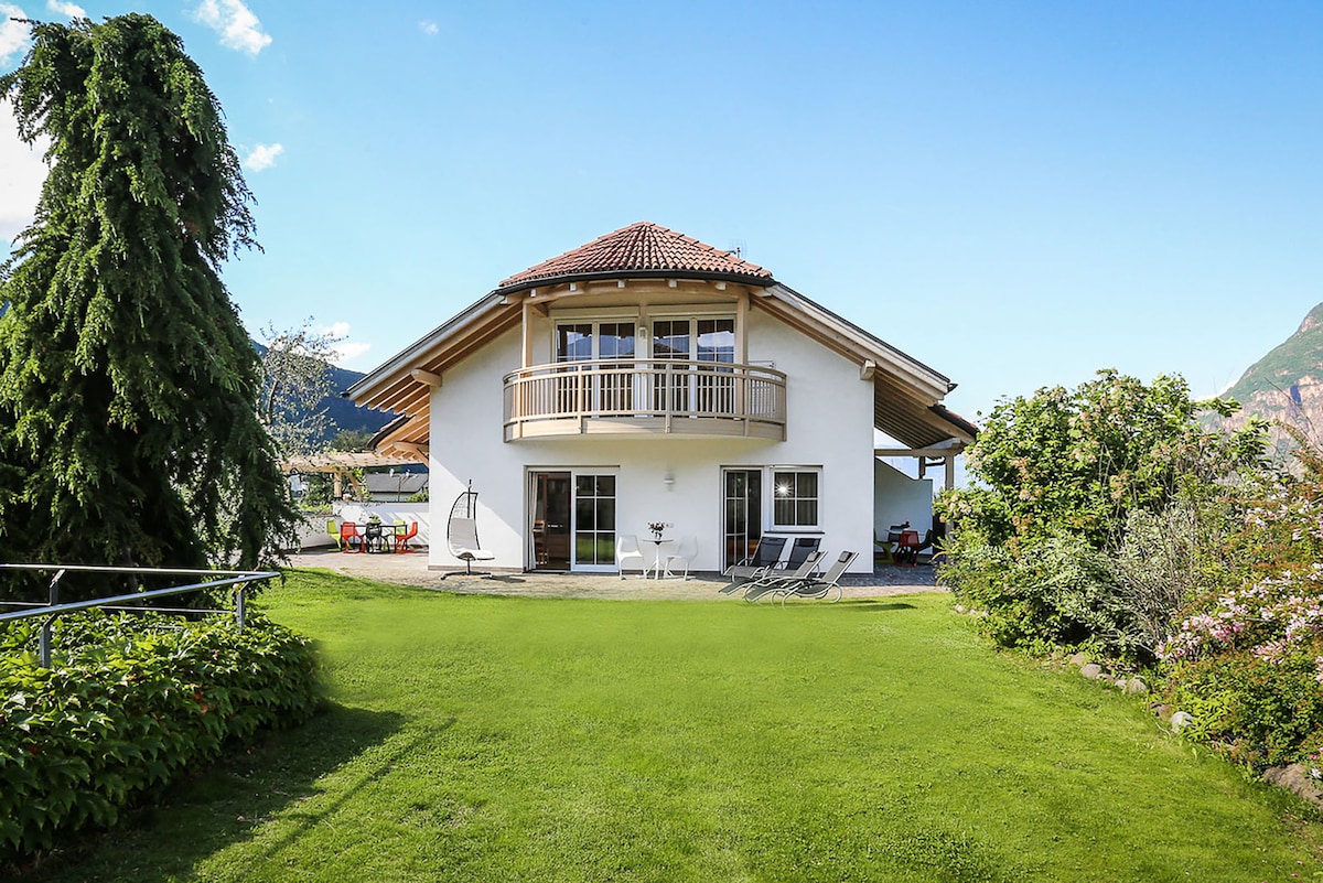 A white villa is situated amidst lush greenery, featuring a spacious lawn and a wraparound balcony. The building showcases a tiled roof and large windows, allowing natural light to fill the interior. Sun loungers and a hanging chair are positioned on the patio, inviting relaxation.