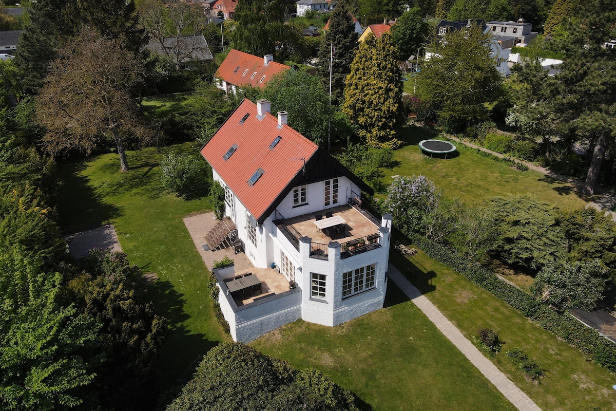 The villa features a striking architectural design, with a distinctive red roof and multiple large windows. The spacious backyard includes a trampoline and lush greenery, providing ample outdoor space for relaxation. A deck is visible, offering areas for seating and enjoying the surrounding nature.