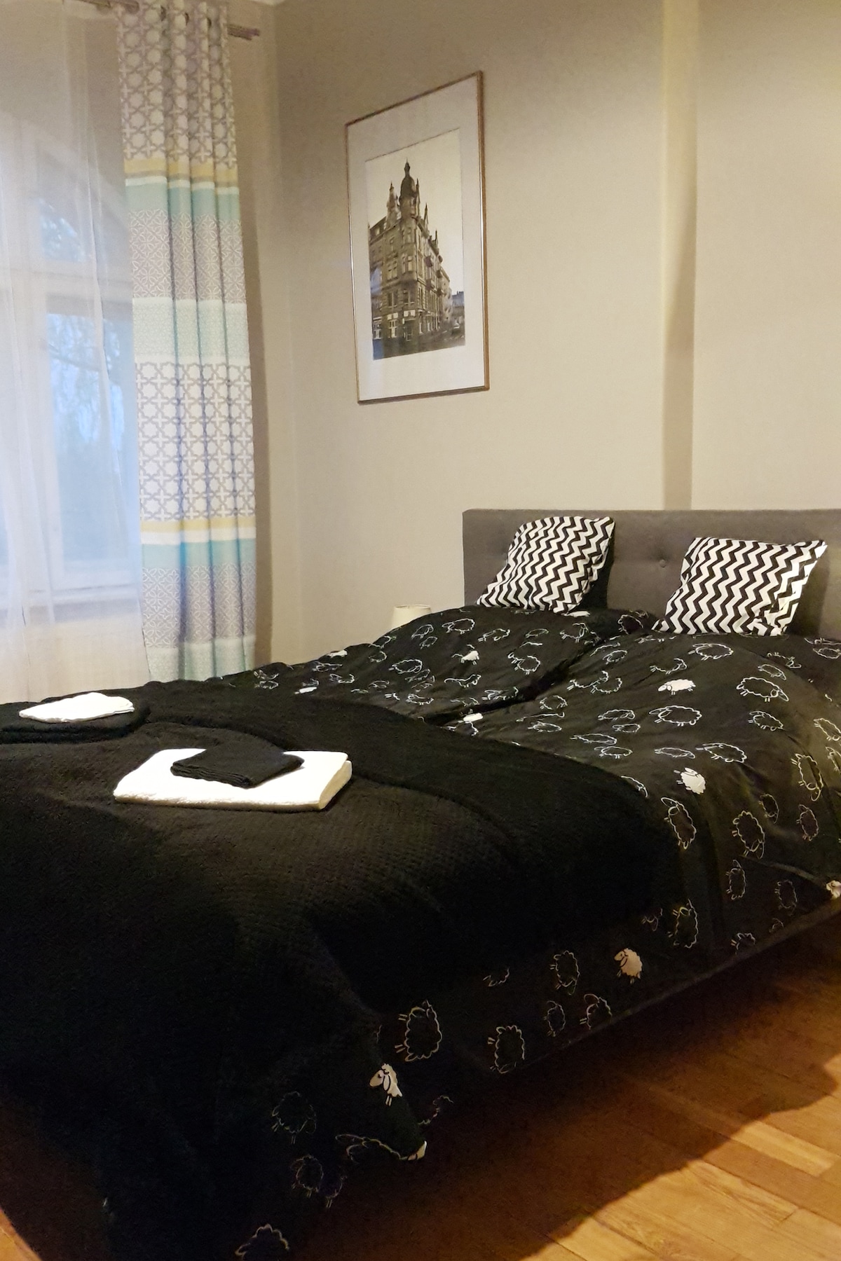 A comfortable sleeping area features a large bed dressed in a dark patterned duvet. Two decorative pillows rest against a headboard. Light-coloured curtains frame the window, while a framed artwork adorns the wall, creating a relaxing ambiance.