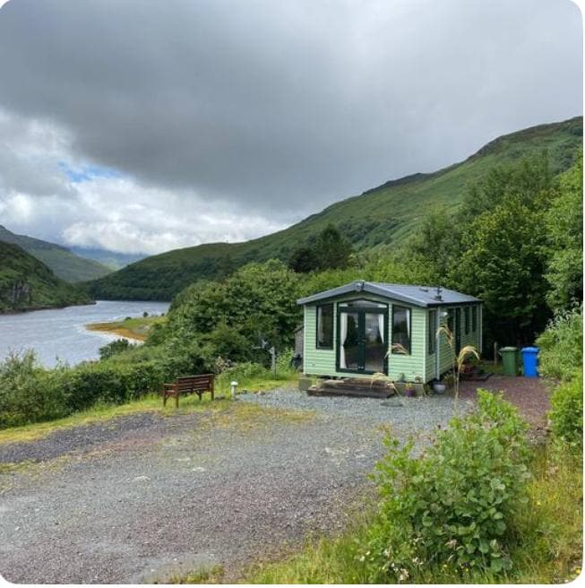 A charming green cabin is situated beside a winding river, framed by rolling hills and lush greenery. A gravel path leads to the entrance, with a wooden bench positioned nearby for seating. The sky is partially cloudy, adding depth to the scene.