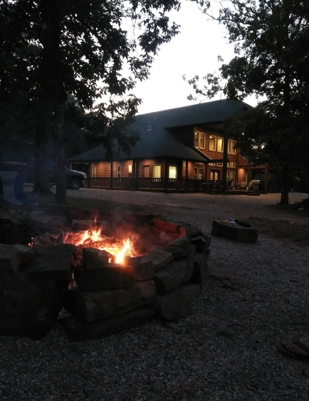 A stone fire pit is prominently featured in the foreground, with flames dancing amid the evening glow. In the background, a spacious cabin is illuminated, showcasing its large windows and inviting porch, surrounded by trees in a serene outdoor setting.