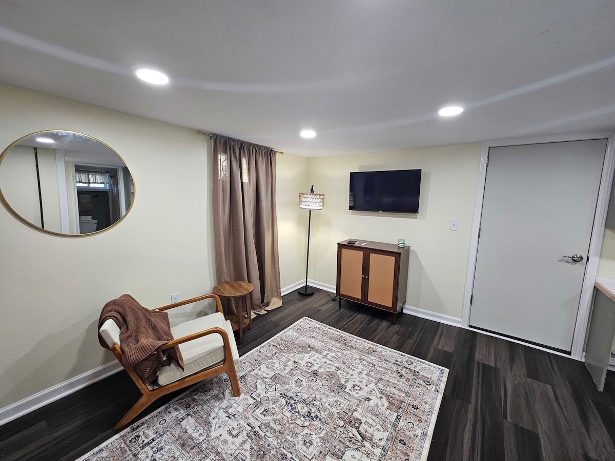 A welcoming living space features a comfortable armchair positioned beside a small accent table. A large mirror and a wall-mounted TV are visible, while a light-colored cabinet provides storage. Soft curtain drapes frame a window, and a patterned area rug adds warmth to the room.