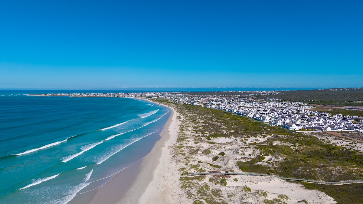A sweeping view showcases the coastline of Langezandt, with gentle waves lapping against a sandy beach. The white cottages of the village are nestled just beyond the shore, framed by lush greenery under a clear blue sky.