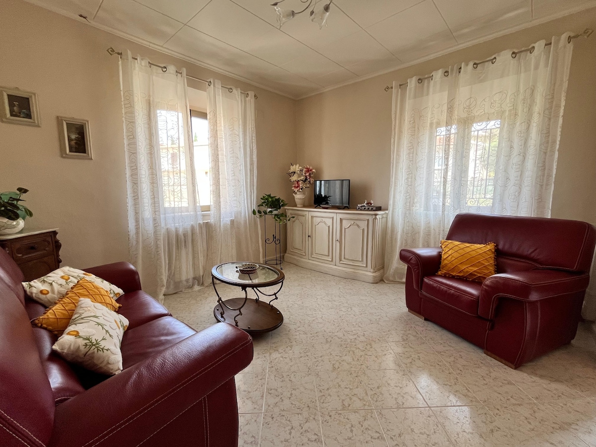 A welcoming living area features two red leather chairs with coordinating decorative cushions. A round glass coffee table occupies the center, while a small television is positioned on a light-colored cabinet. Natural light enters through two windows dressed in sheer white curtains.