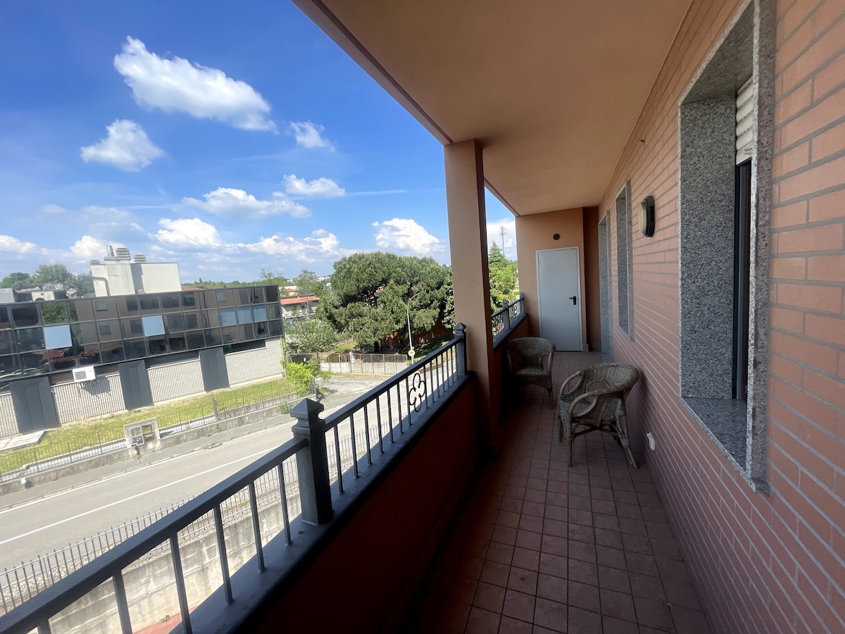 A spacious balcony is visible, featuring two comfortable wicker chairs. The view includes greenery and a street below, framed by a brick facade. The open sky above displays a few scattered clouds, contributing to a light and airy atmosphere.
