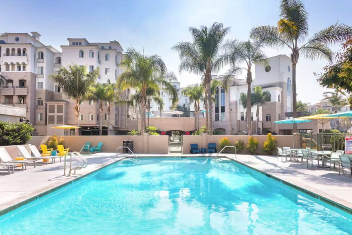 A serene pool area is surrounded by lush palm trees, with lounge chairs positioned beside the water. Sun umbrellas in various colors provide shade, while the modern architecture of the surrounding buildings enhances the inviting atmosphere. The pool's clear water reflects the bright sky.