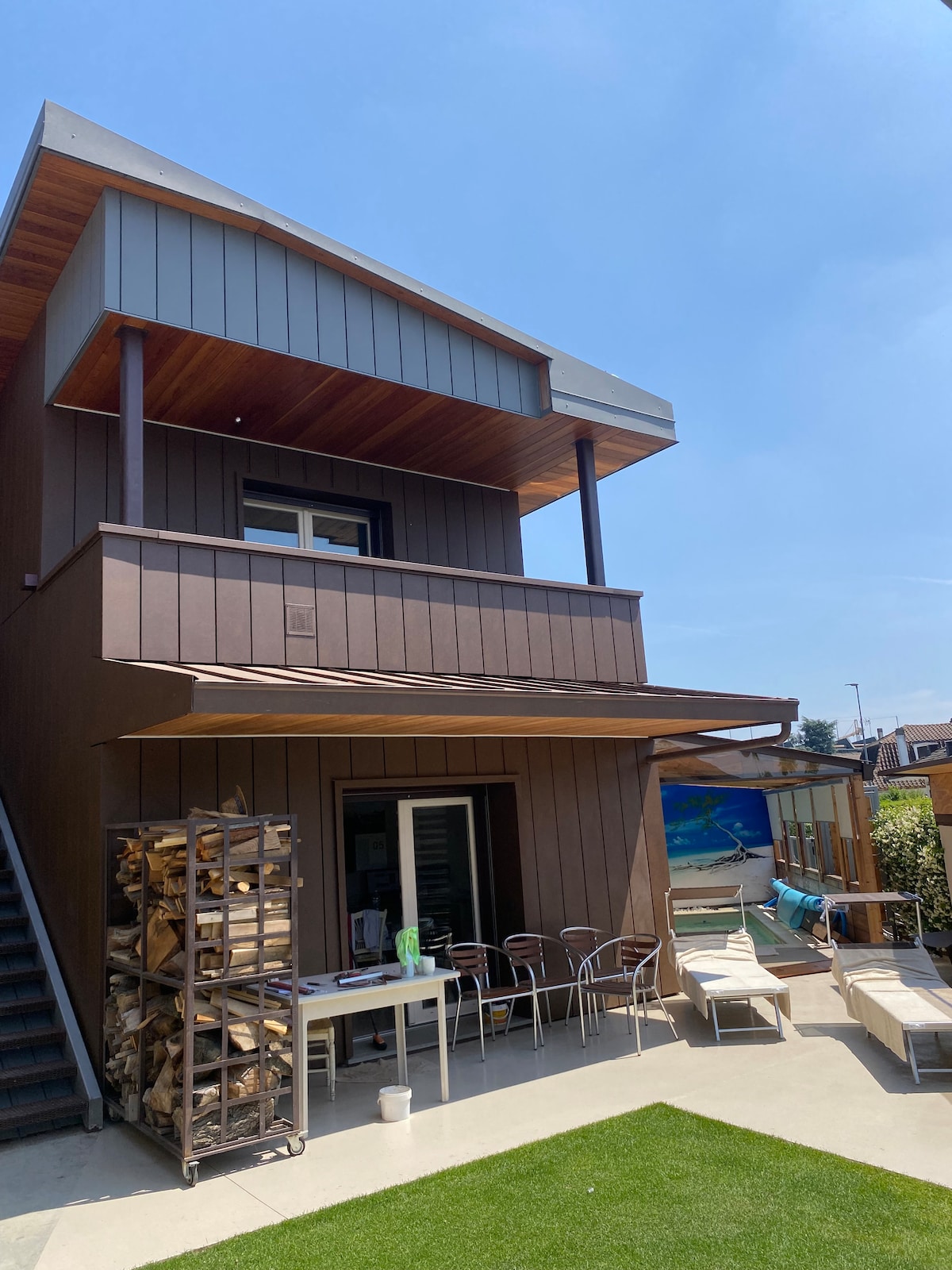 An exterior view of a modern building is captured, showcasing a two-story structure. A spacious outdoor area is visible, featuring stacked wood, a dining table with several chairs, and sun loungers. The clear blue sky provides a bright backdrop to the well-maintained landscape.