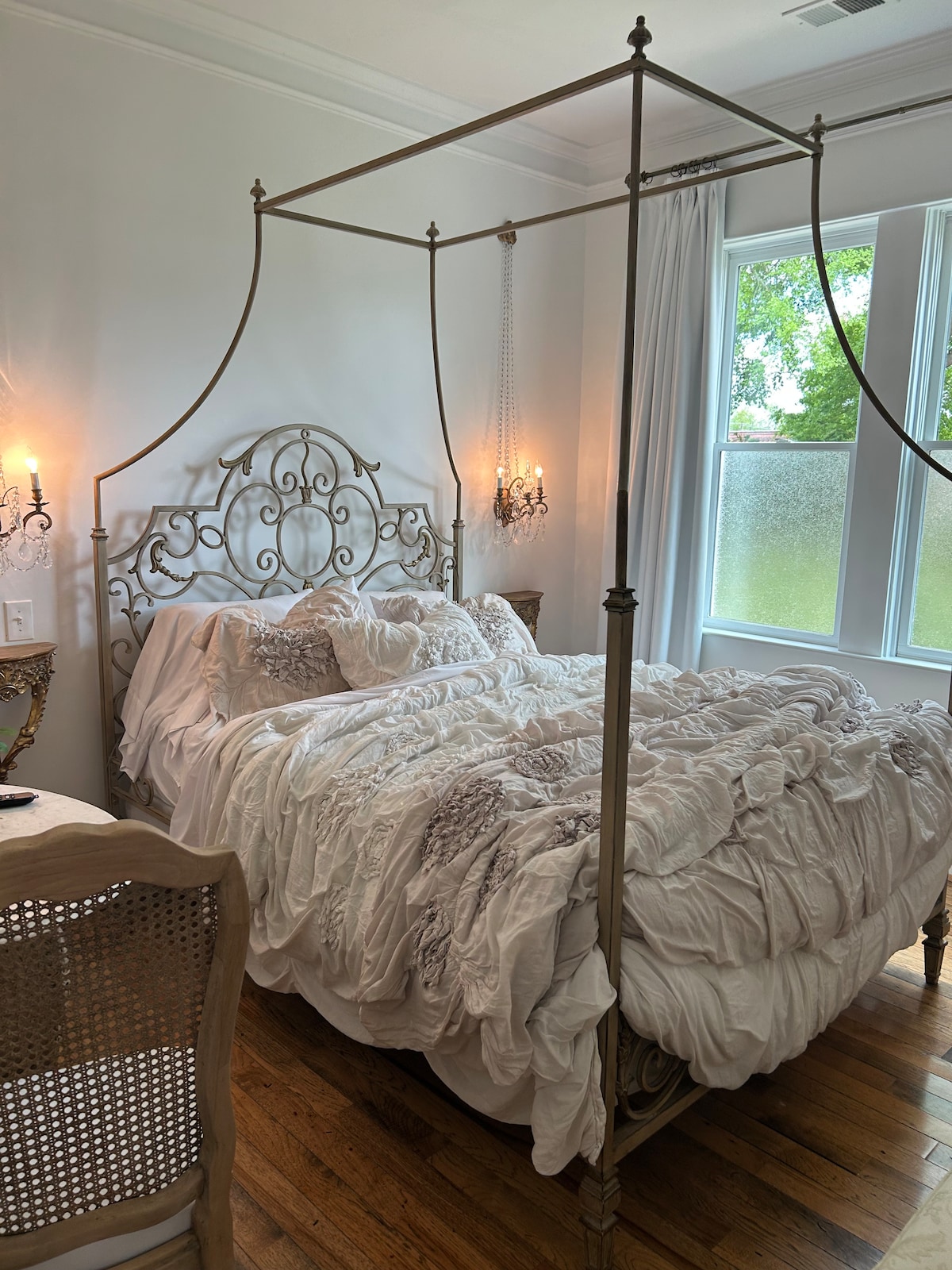 A spacious bed with a decorative metal canopy frame is highlighted in the room. Soft bedding in light tones is neatly arranged. Natural light enters through large windows, while textured curtains add privacy. Elegant sconces provide ambient lighting on either side of the bed.