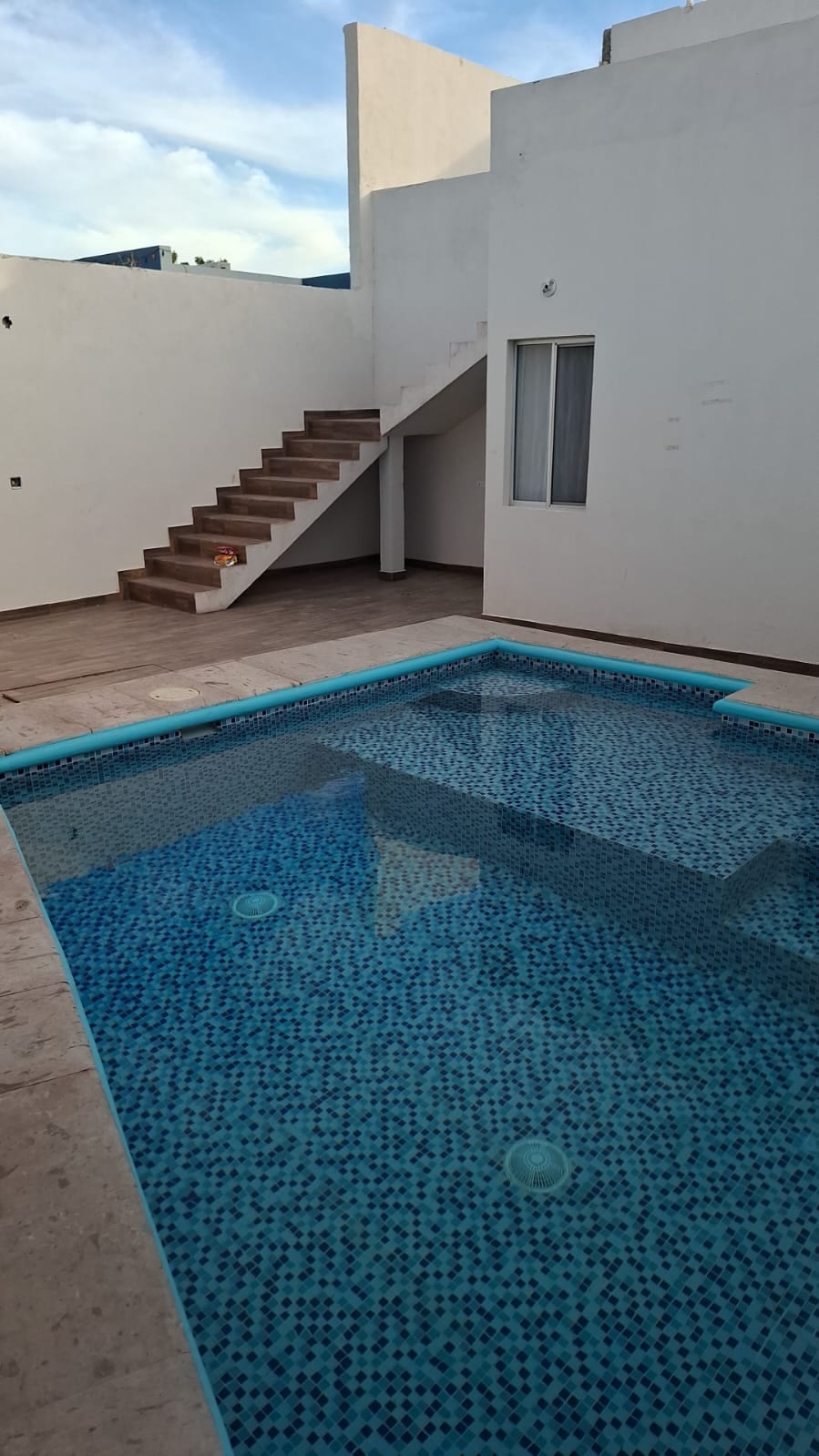 A private pool is situated in a well-lit outdoor area, surrounded by smooth tiled flooring. A staircase is visible in the background, leading to an upper level of the property. The walls are painted in a clean white hue, enhancing the open atmosphere.